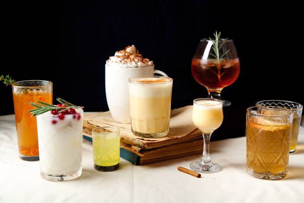 Various colorful cocktails on a table, including eggnog, with garnishes, next to an old book, dark background.