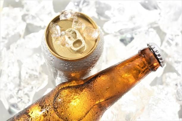 A beer bottle and a can in ice, with water droplets on the cold surfaces.