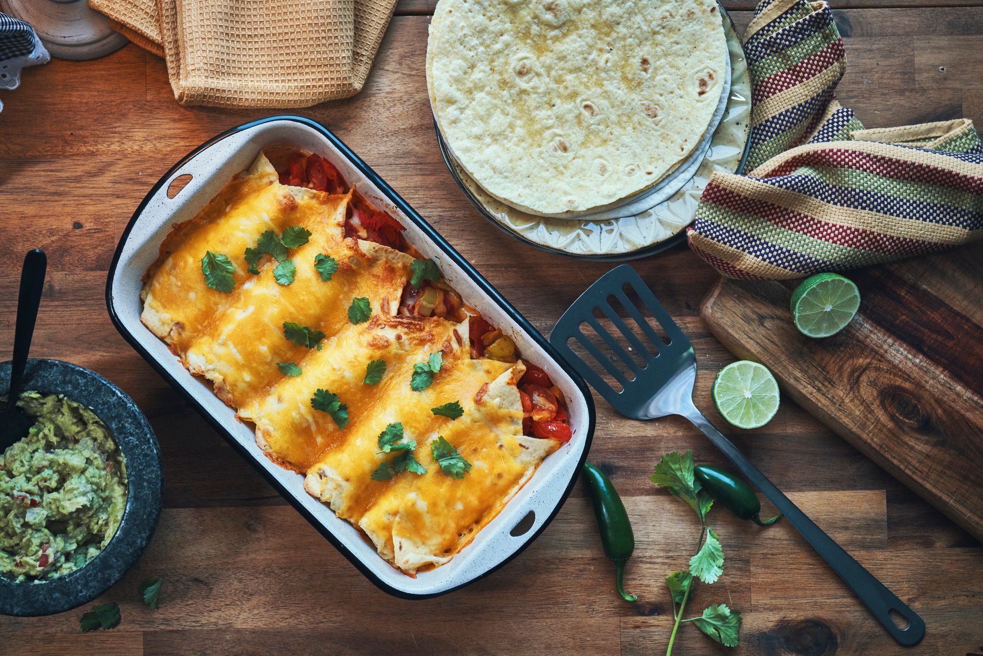 Enchiladas in a baking dish, with guacamole, tortillas, limes, and jalapenos on a wooden table. Enchiladas in a baking dish, with guacamole, tortillas, limes, and jalapenos on a wooden table.