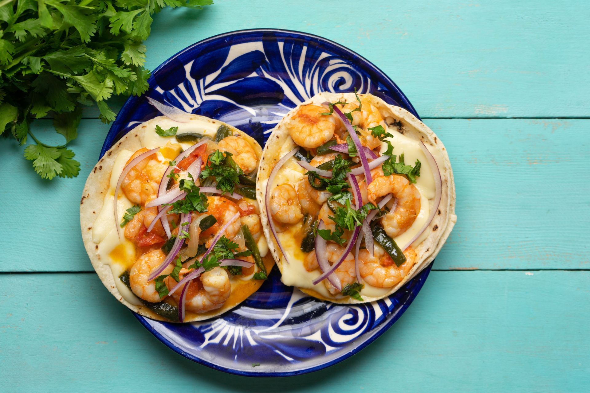 Shrimp tacos on blue plate, topped with onions, cilantro, and melted cheese, set against a teal background. Shrimp tacos on blue plate, topped with onions, cilantro, and melted cheese, set against a teal background.