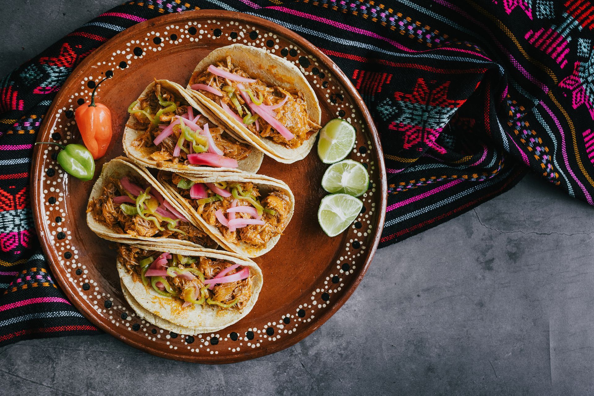 Tacos on a brown plate with habanero pepper, lime wedges, and a colorful textile background.