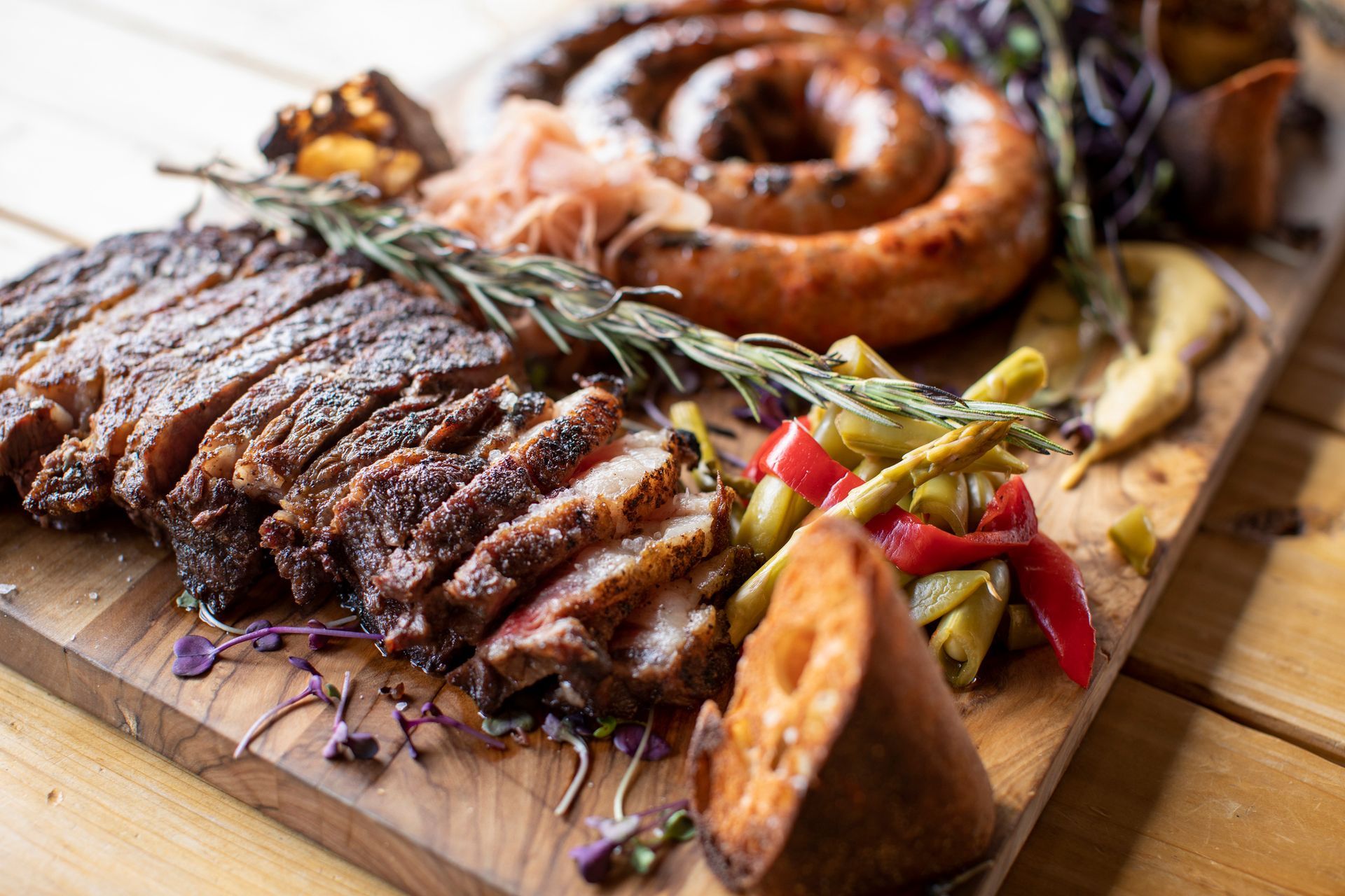 Sliced grilled steak, coiled sausage, vegetables, and sauce on a wooden serving board. Sliced grilled steak, coiled sausage, vegetables, and sauce on a wooden serving board.