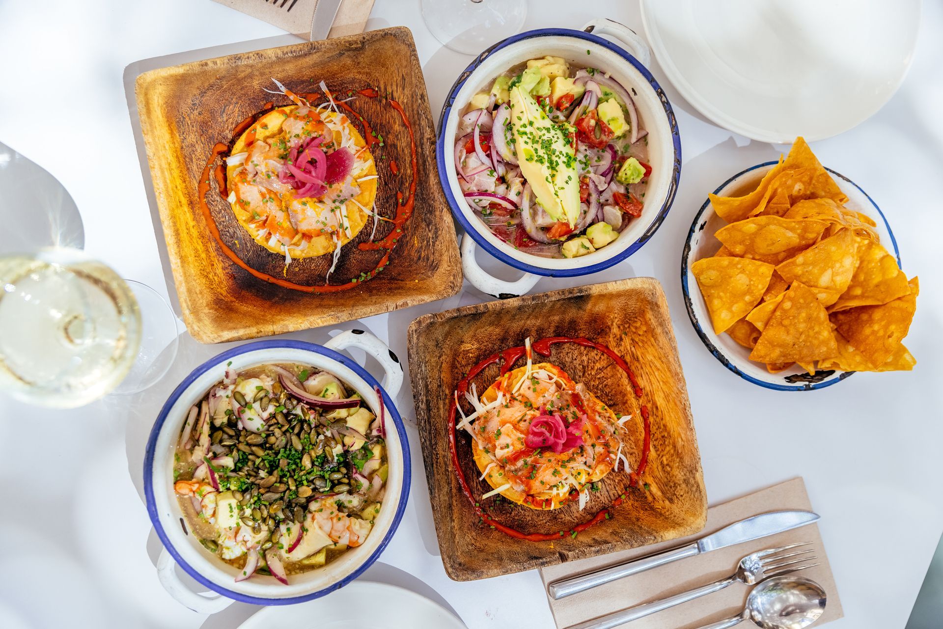 Overhead shot of a table with tostadas, ceviche bowls, tortilla chips, and a glass of wine.