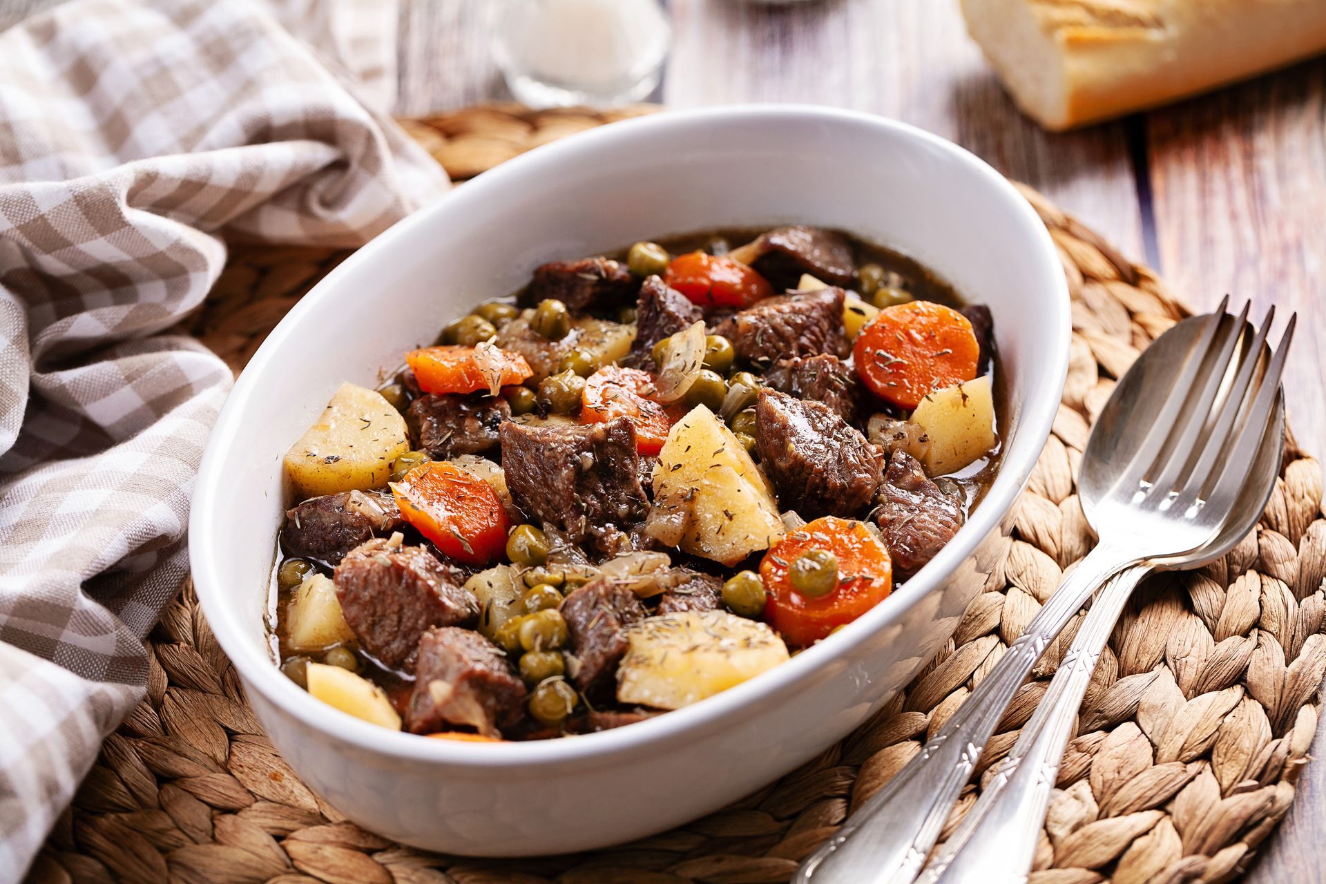 Beef stew in a white bowl with carrots, potatoes, and peas; fork and bread in the background. Beef stew in a white bowl with carrots, potatoes, and peas; fork and bread in the background.
