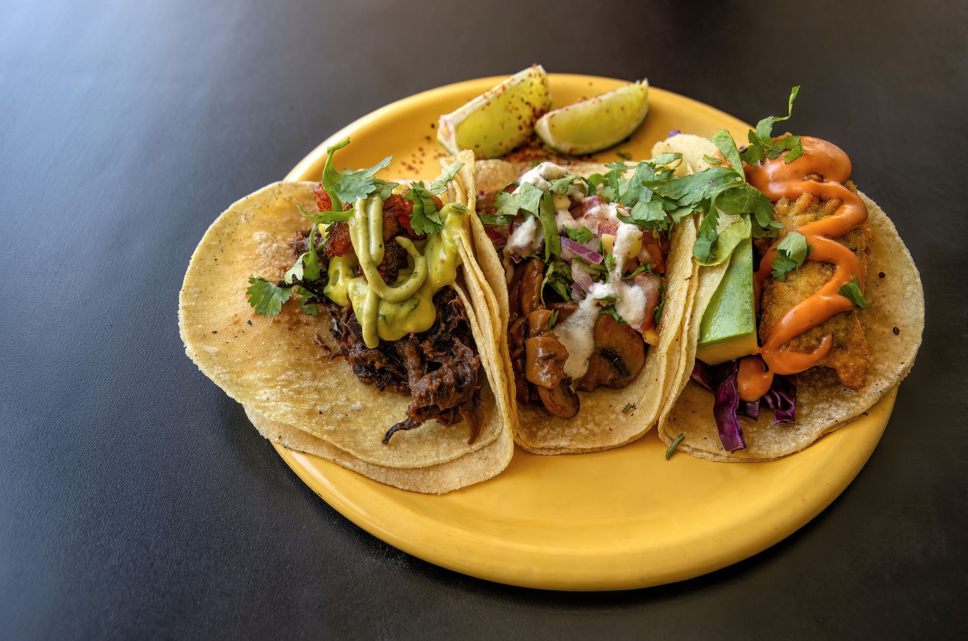 Three tacos on a yellow plate with lime wedges, cilantro, and various toppings.