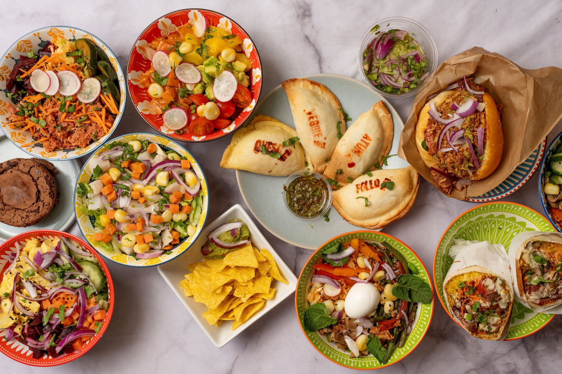 Assorted dishes including salads, empanadas, and a burrito on a marble surface. Assorted dishes including salads, empanadas, and a burrito on a marble surface.