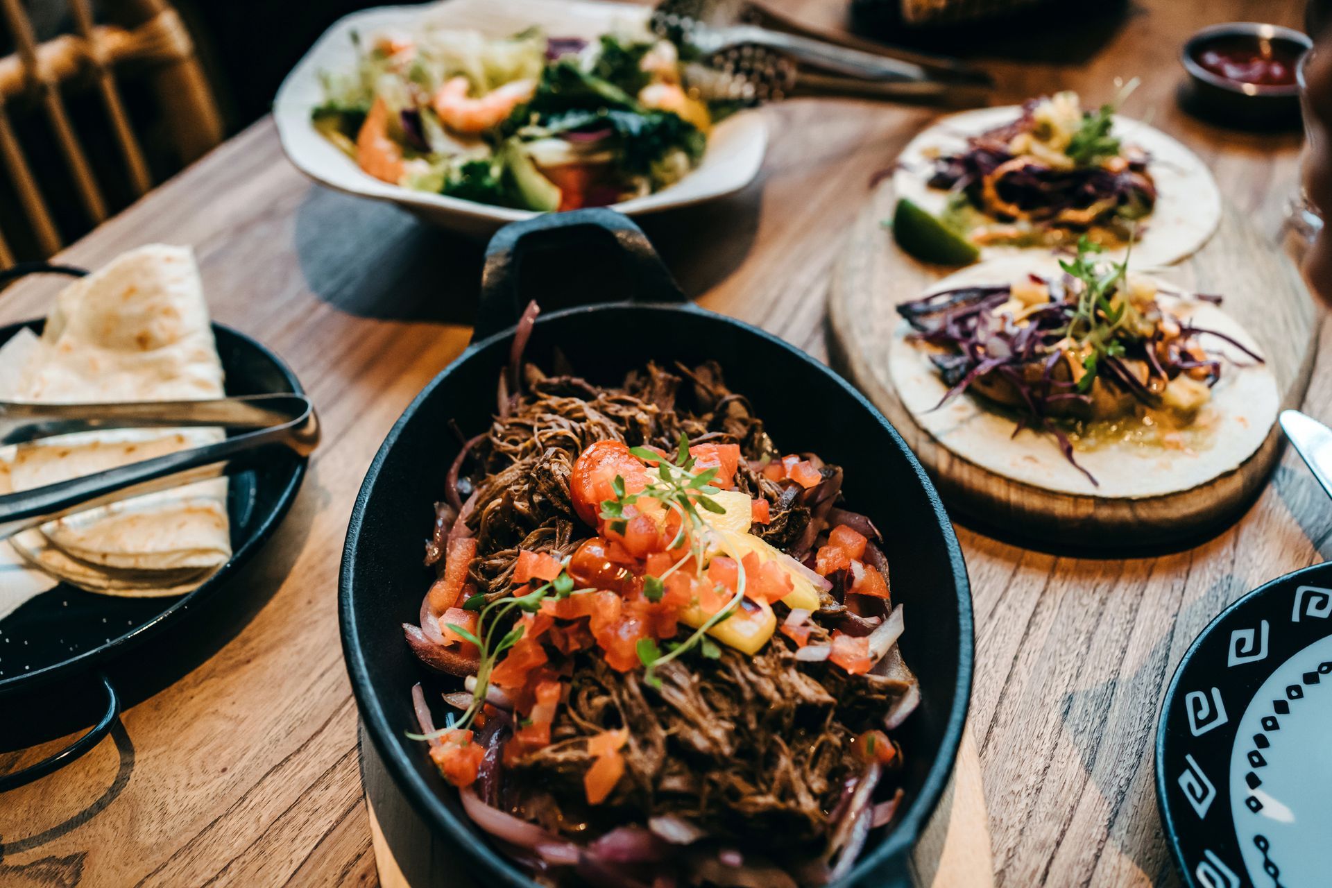 Table with assorted food: tacos, salad, meat dish, tortillas. Wooden surface, neutral lighting. Table with assorted food: tacos, salad, meat dish, tortillas. Wooden surface, neutral lighting.