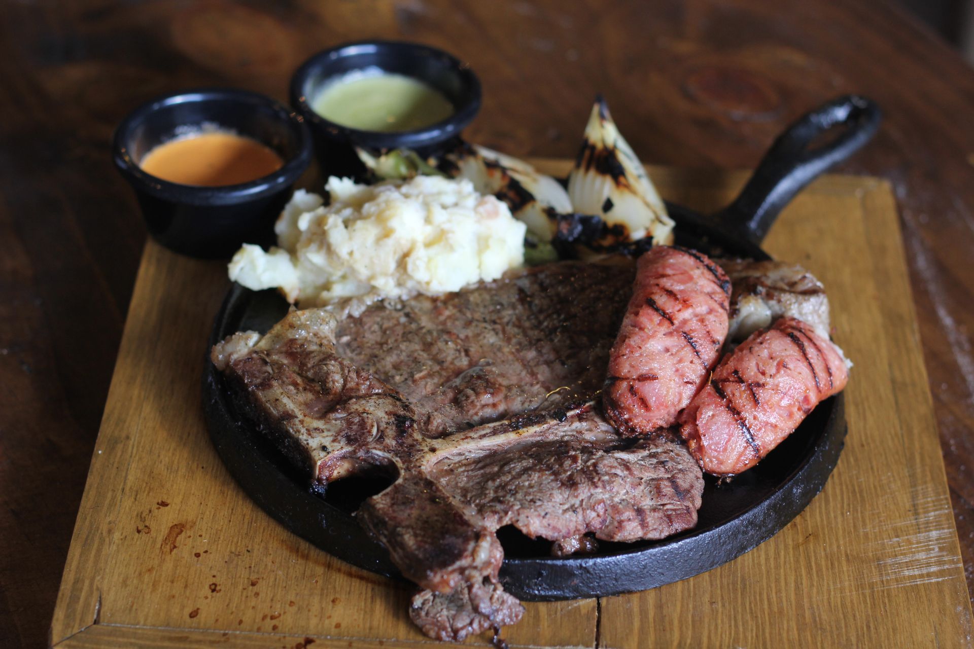 T-bone steak with grilled sausage, mashed potatoes, and sauces on a cast iron skillet, served on a wooden board. T-bone steak with grilled sausage, mashed potatoes, and sauces on a cast iron skillet, served on a wooden board.