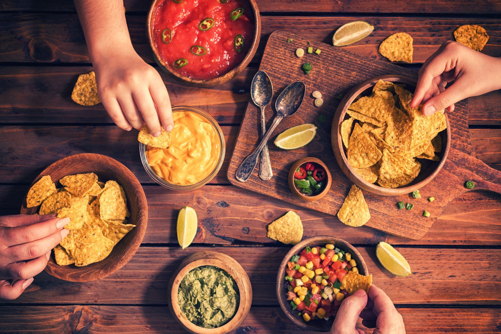 People dipping tortilla chips into various salsas and dips on a wooden table. People dipping tortilla chips into various salsas and dips on a wooden table.