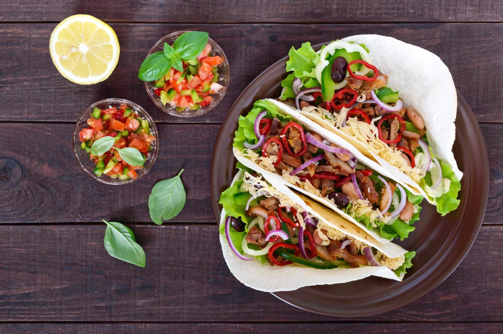 Tacos with toppings on a brown plate, served with salsa and lemon on a wooden table.