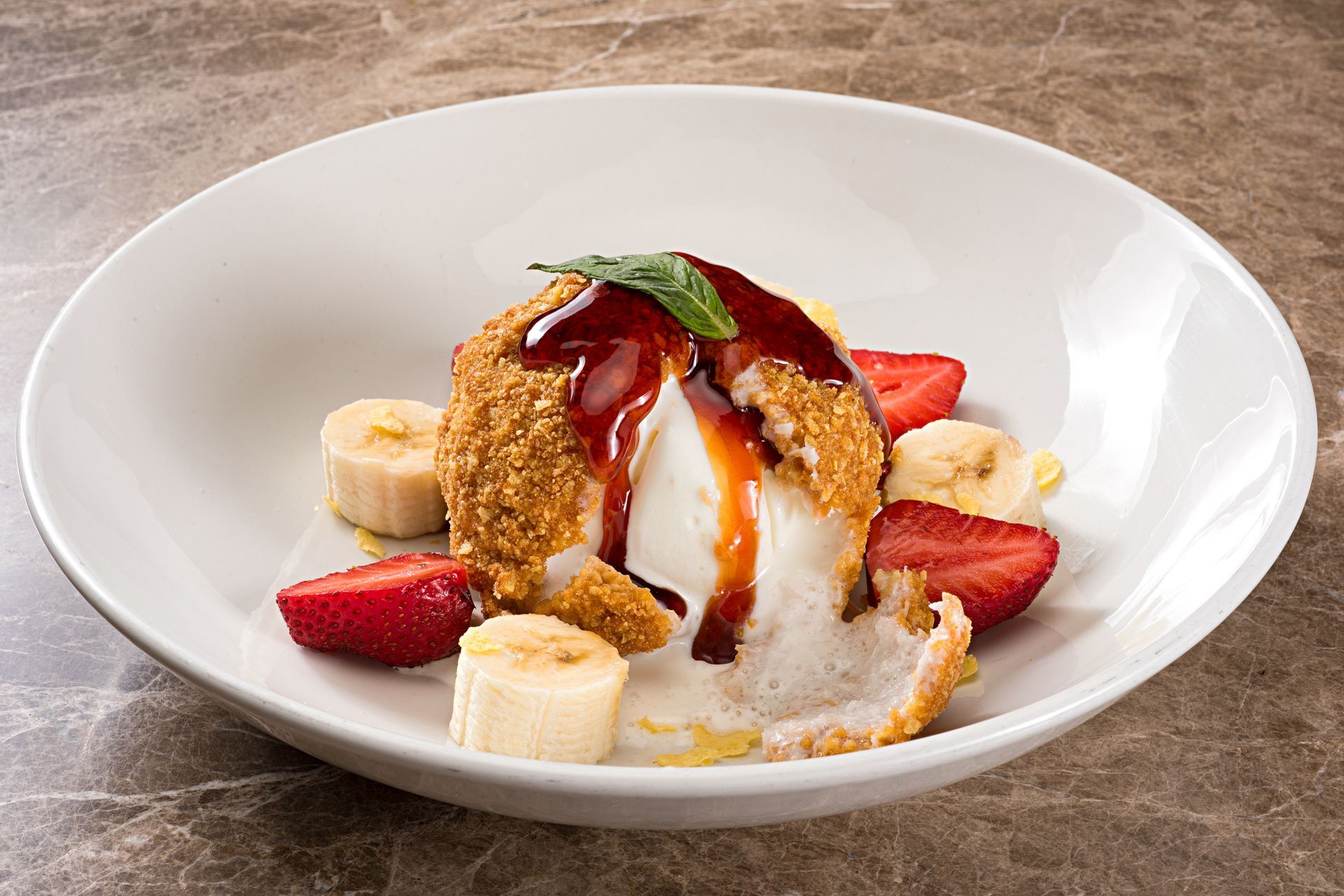 Fried ice cream dessert with strawberries and bananas, drizzled with sauce in a white bowl.