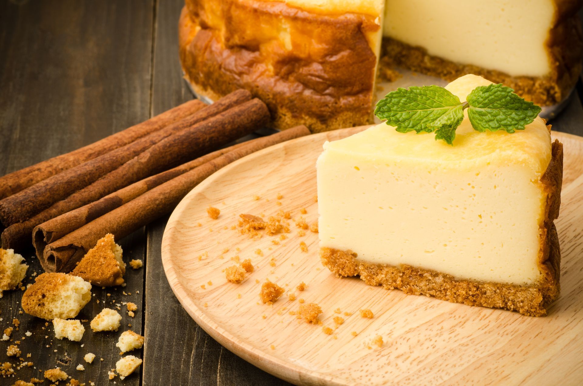 Cheesecake slice with mint garnish on a wooden plate, cinnamon sticks, and crumbs. Another cheesecake in the background.