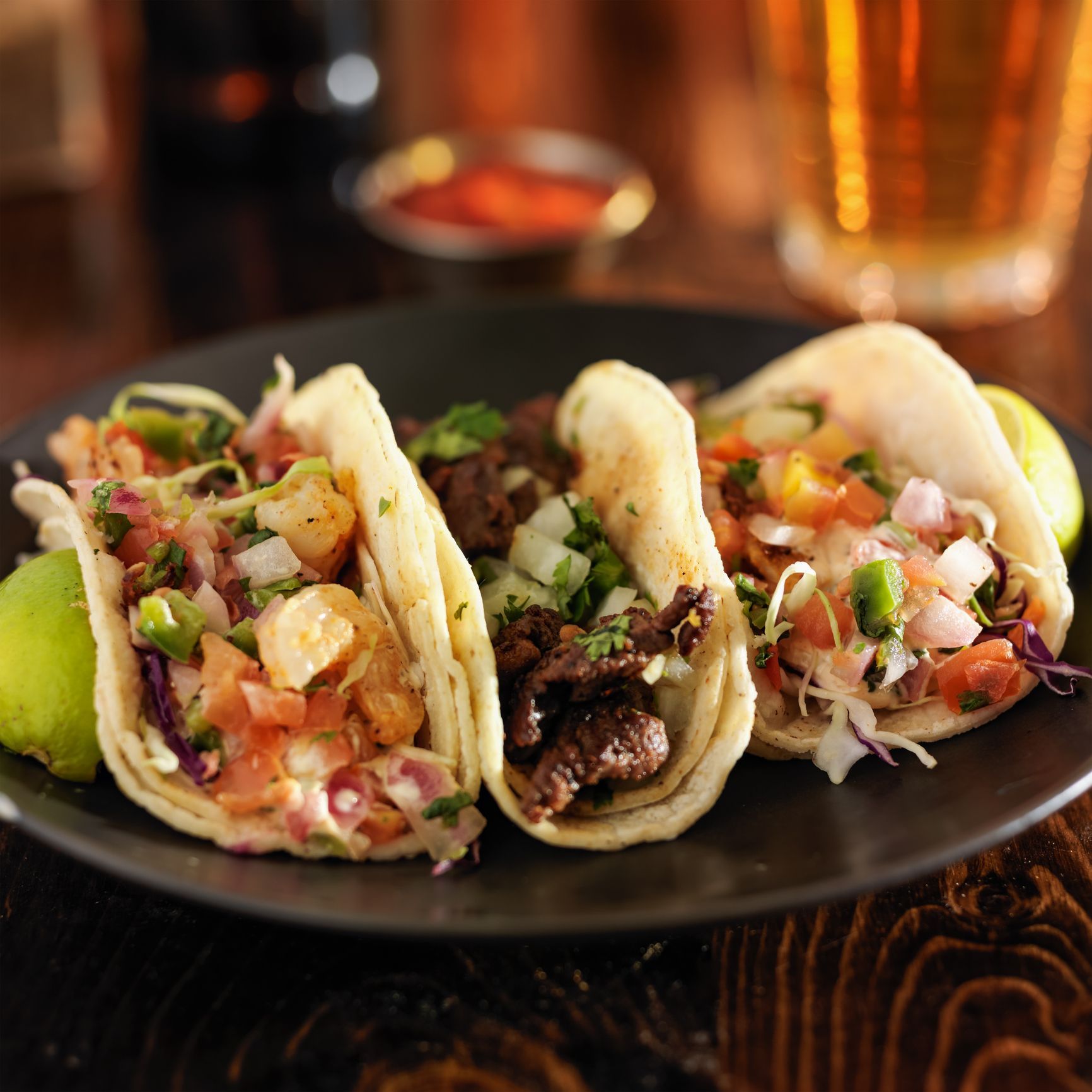 Three tacos on a black plate with shrimp, steak, and salsa; a glass of beer in the background.