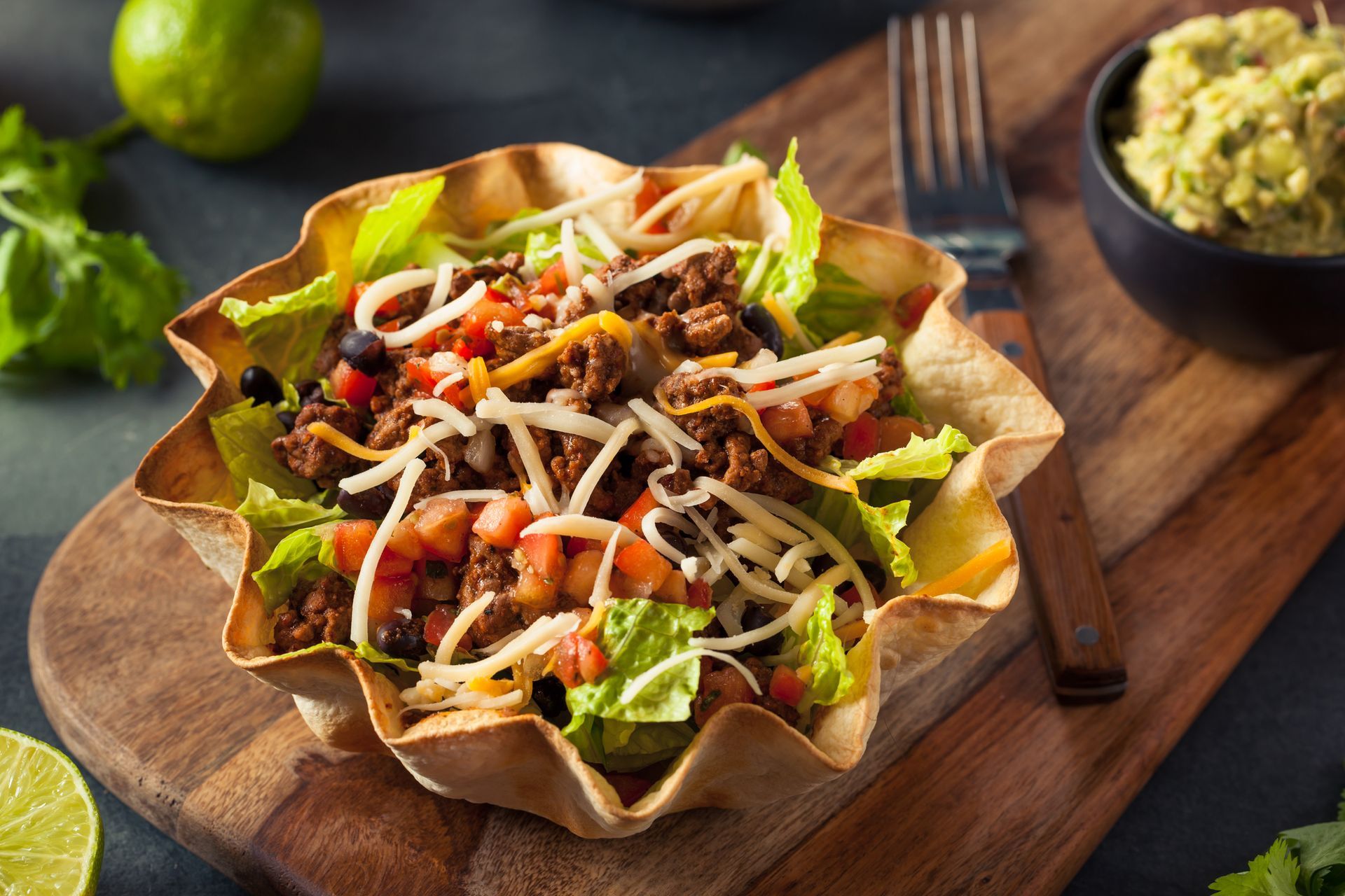 Taco salad in a crispy shell with meat, cheese, tomatoes, and lettuce, alongside guacamole and a lime.