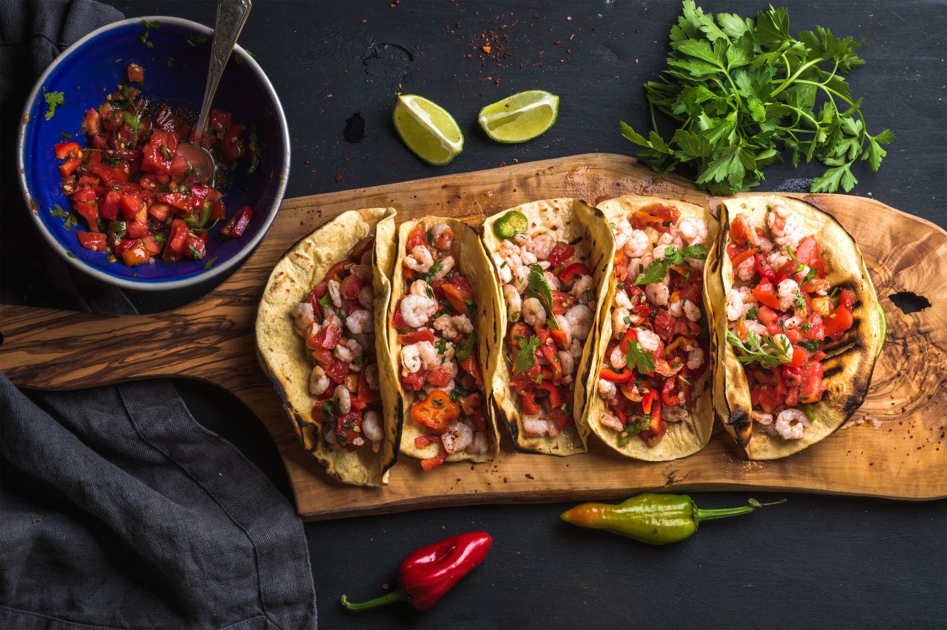 Shrimp tacos with salsa on a wooden board; limes, peppers, and parsley.