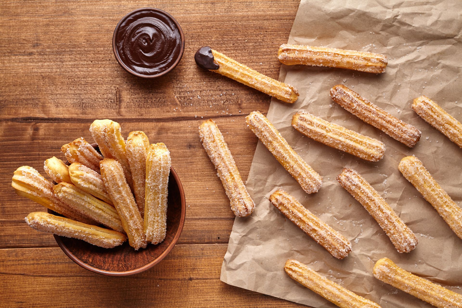 Churros with chocolate dip on a wooden table. Some churros are arranged on parchment paper, others in a wooden bowl.