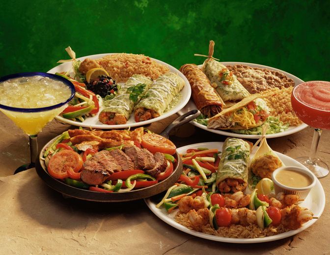 Platters of Mexican food, including fajitas, enchiladas, and tacos, alongside margaritas on a table.
