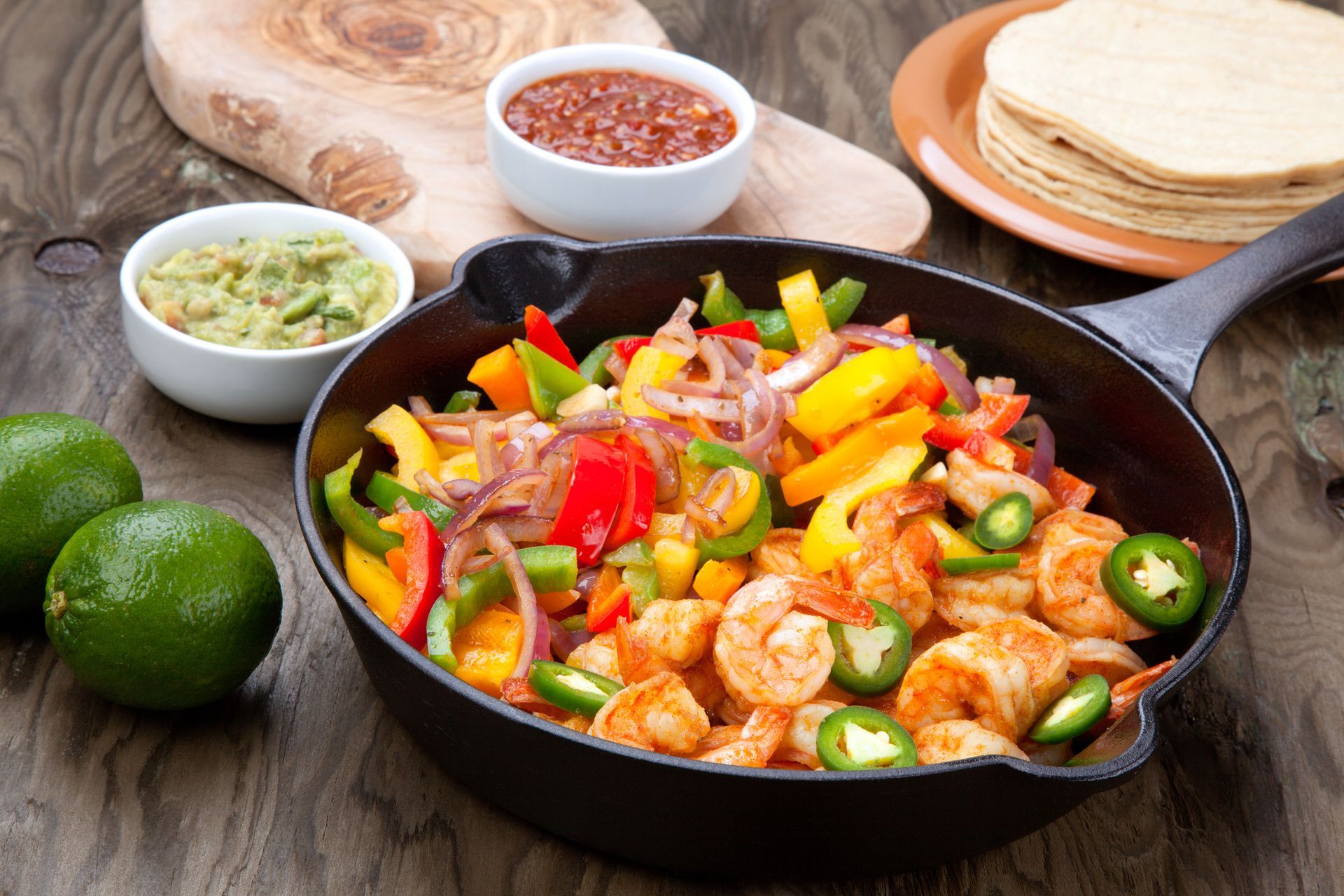 Shrimp fajitas in a cast-iron skillet with colorful peppers, onions, and jalapenos, served with salsa, guacamole, tortillas, and limes.