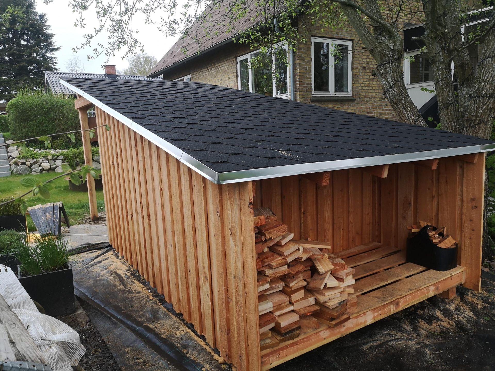 Et træskur med sort tag er fyldt med træstammer.