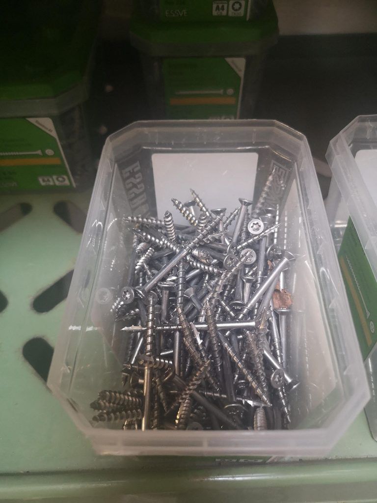 En plastikbeholder fyldt med skruer står på en hylde.
