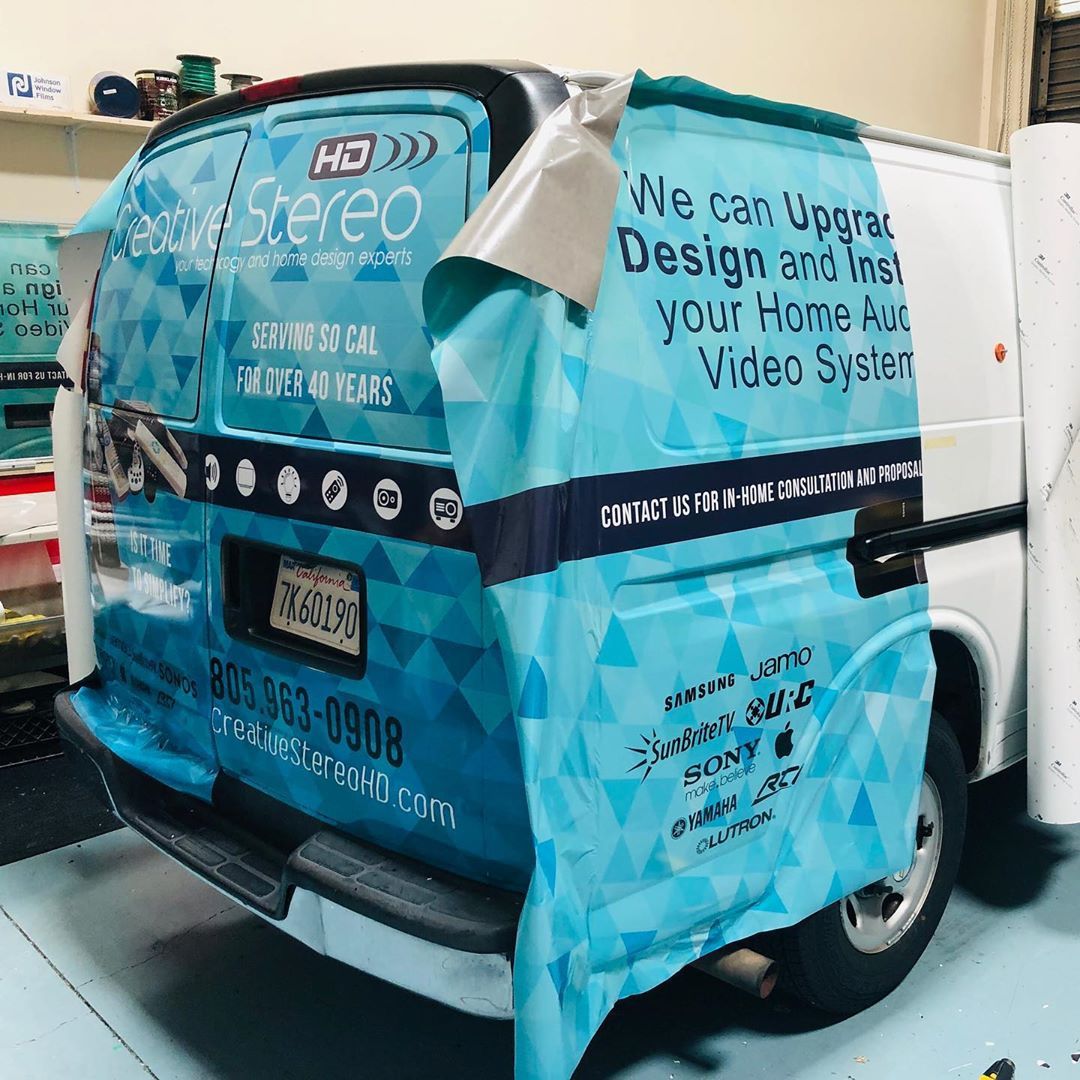 A van is wrapped in a blue and white graphic that says we can upgrade design and install your home audio video system