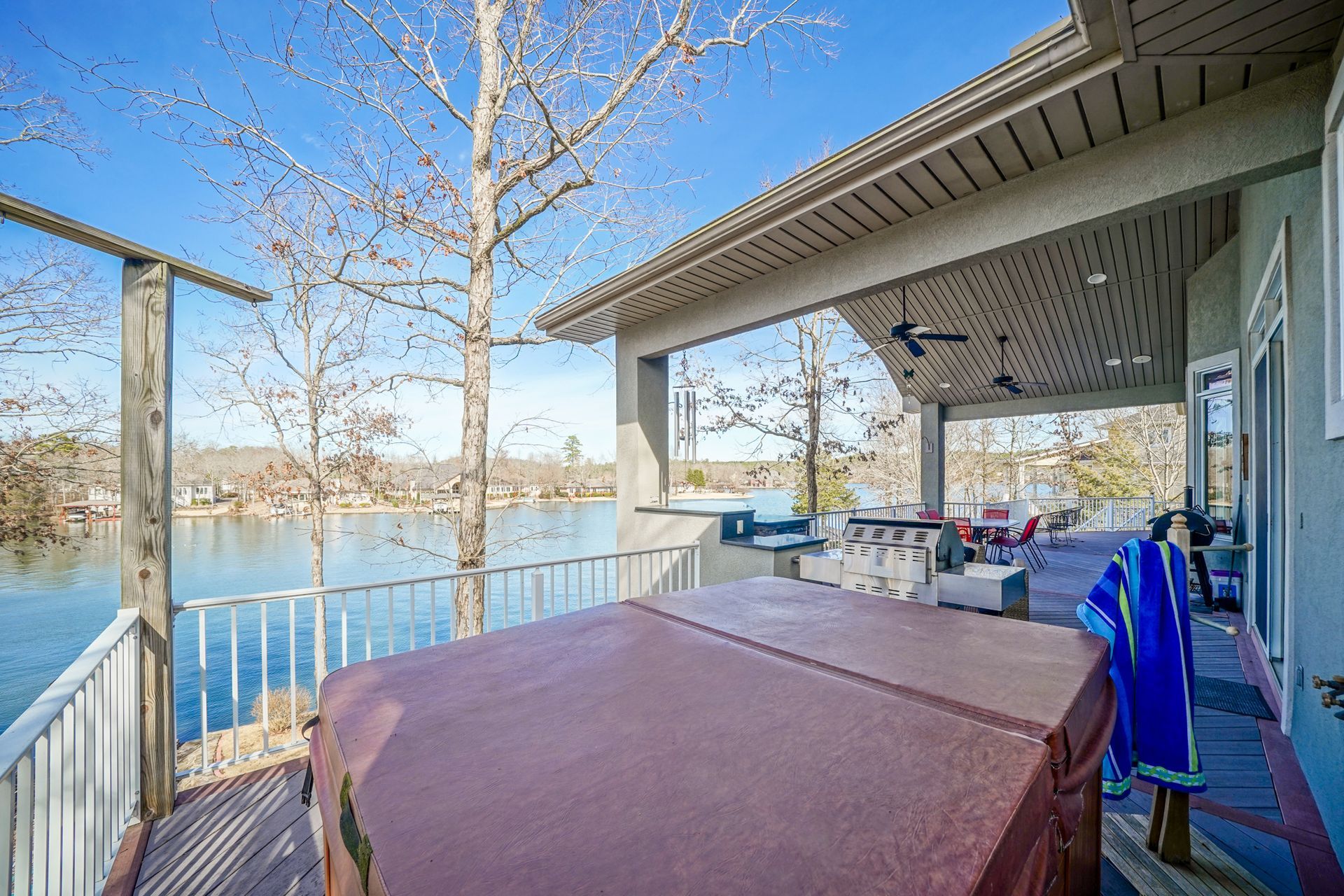 Deck with hot tub overlooking a lake. Trees, white railing, and covered patio area visible.