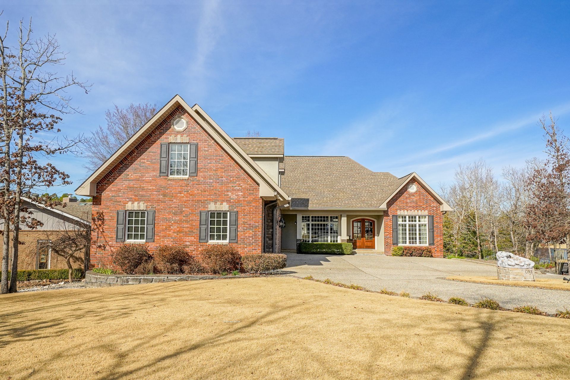 Red brick house with brown roof and shutters, gravel driveway, and dry grass lawn under a blue sky.