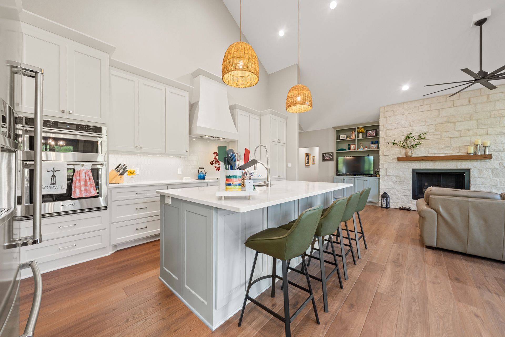 A kitchen with white cabinets , a large island , stools and a ceiling fan.