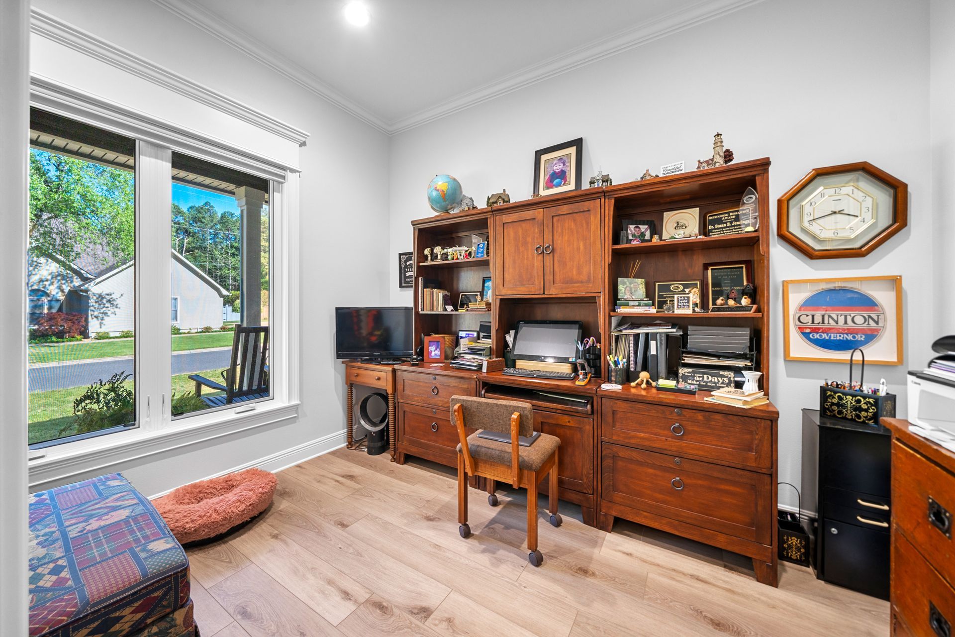 A room with a desk , chair , dresser , and a large window.