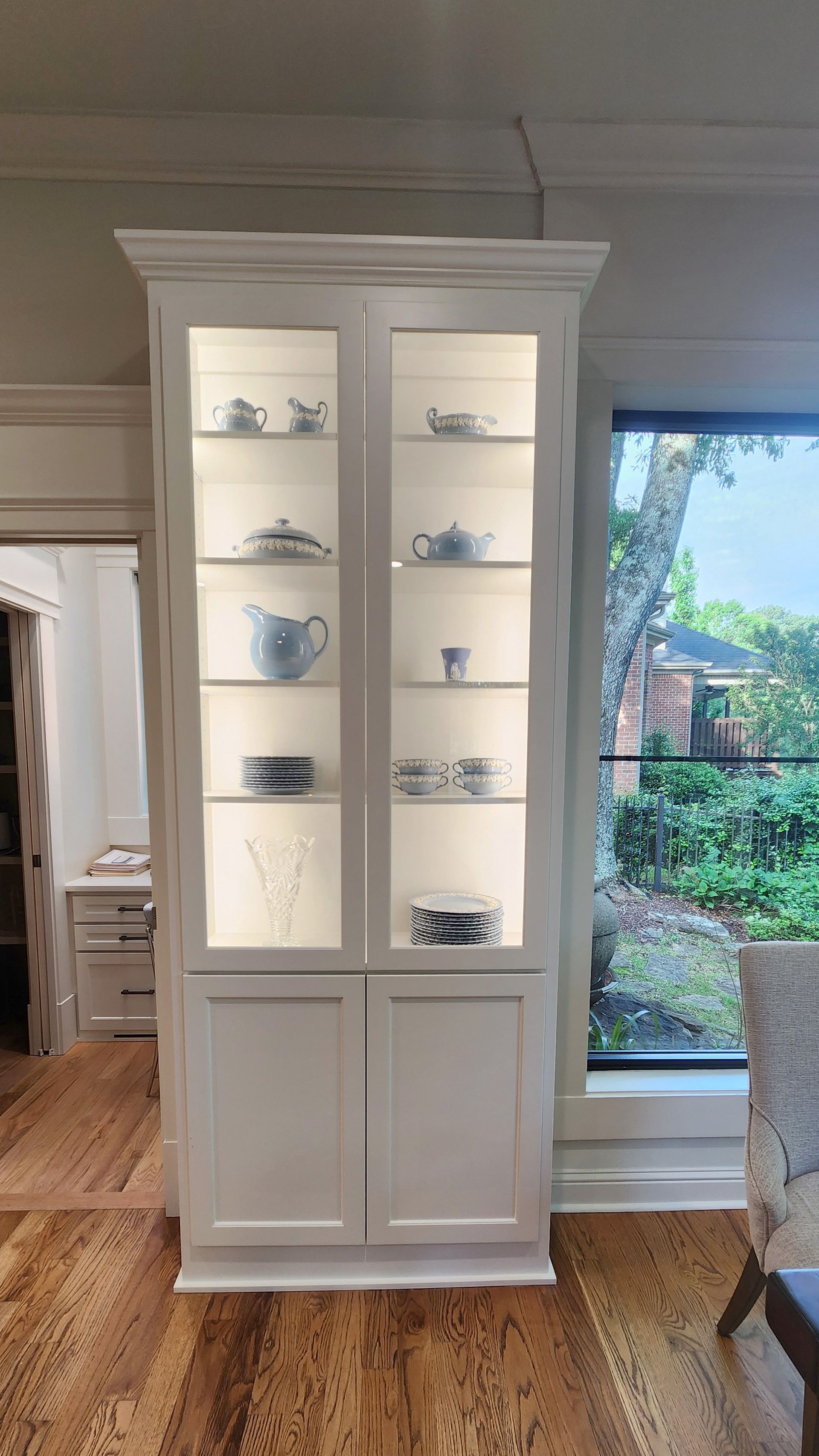 A white cabinet with glass doors and shelves in a living room next to a window.