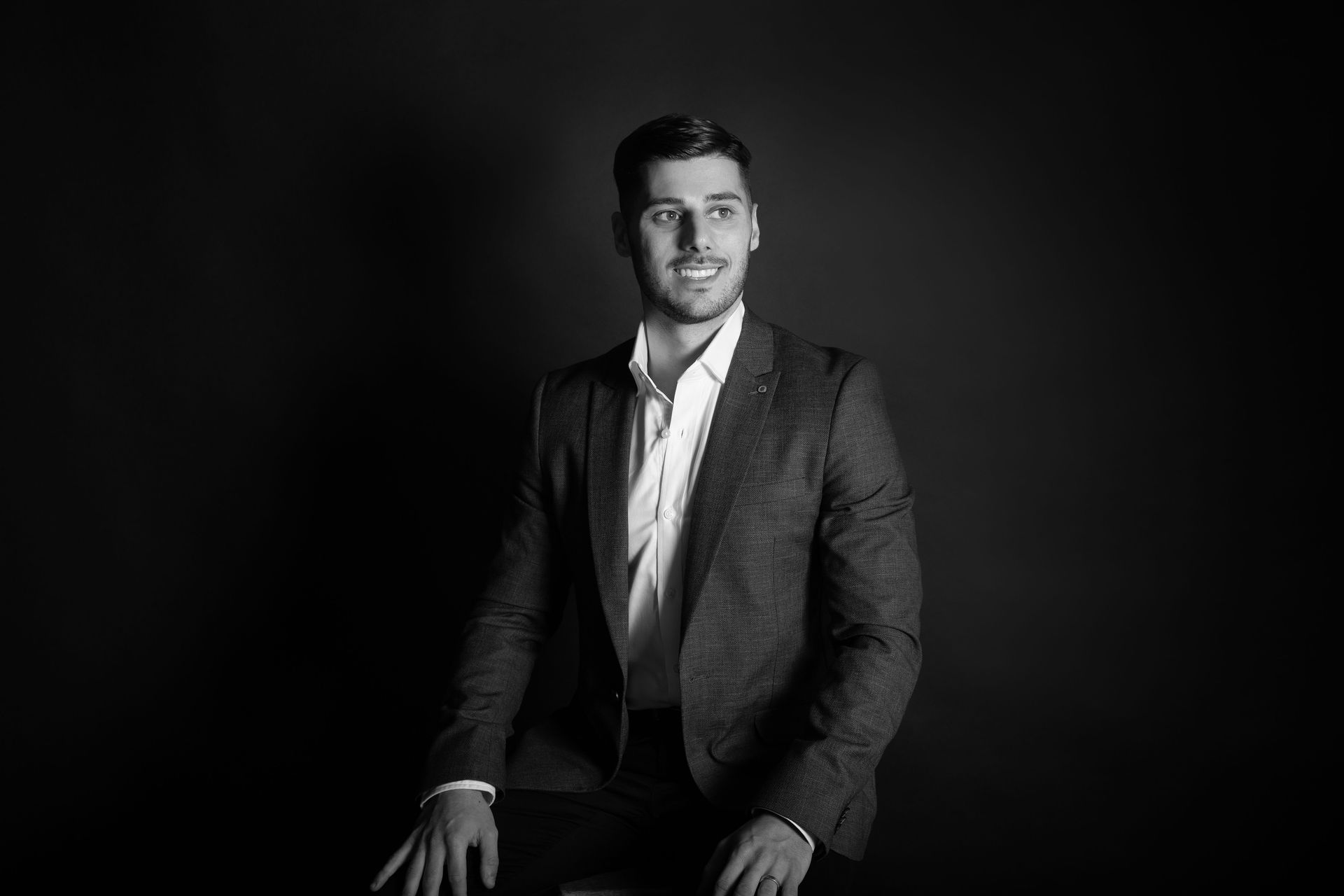 A man in a suit smiles, looking off-camera against a dark background.