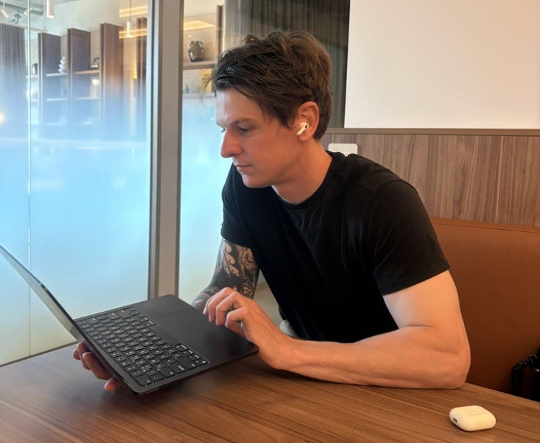 Man wearing AirPods, typing on a laptop, at a table near a window. He wears a black t-shirt and has a tattoo.