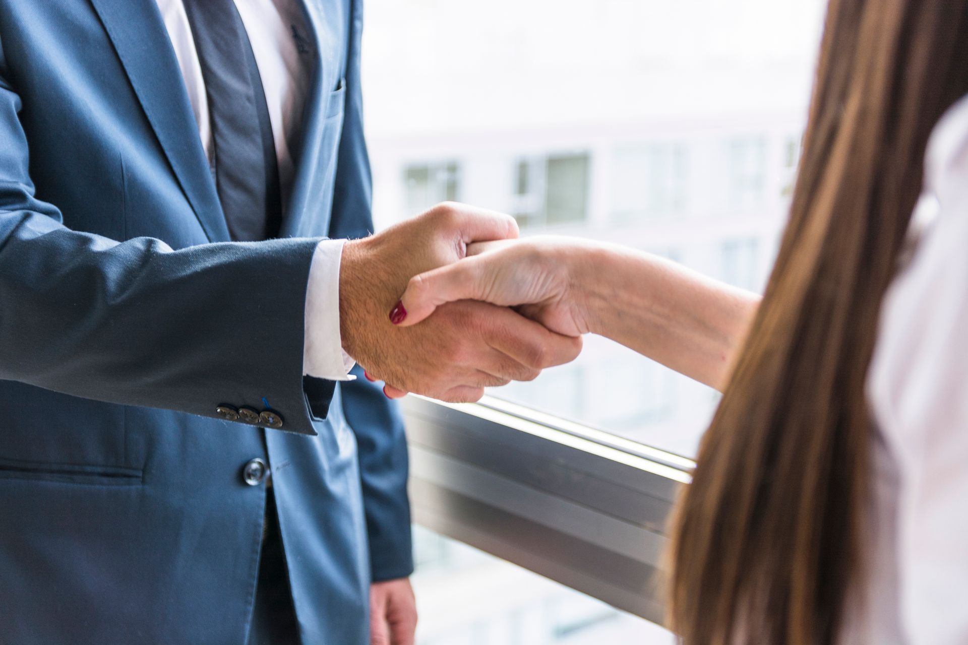 Man in a suit shaking hands with a woman with long brown hair, in front of a window.