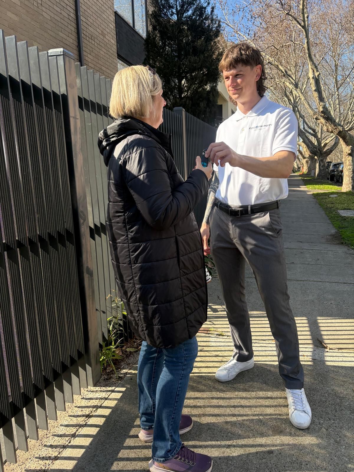 Woman receiving keys from a man outside. The man wears a white polo and gray pants. The woman is in a black coat and jeans.