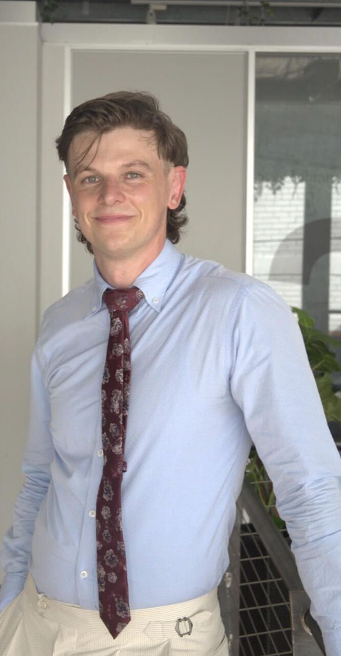 Man with a mullet and tie smiles outdoors, wearing a light blue shirt and khaki pants.