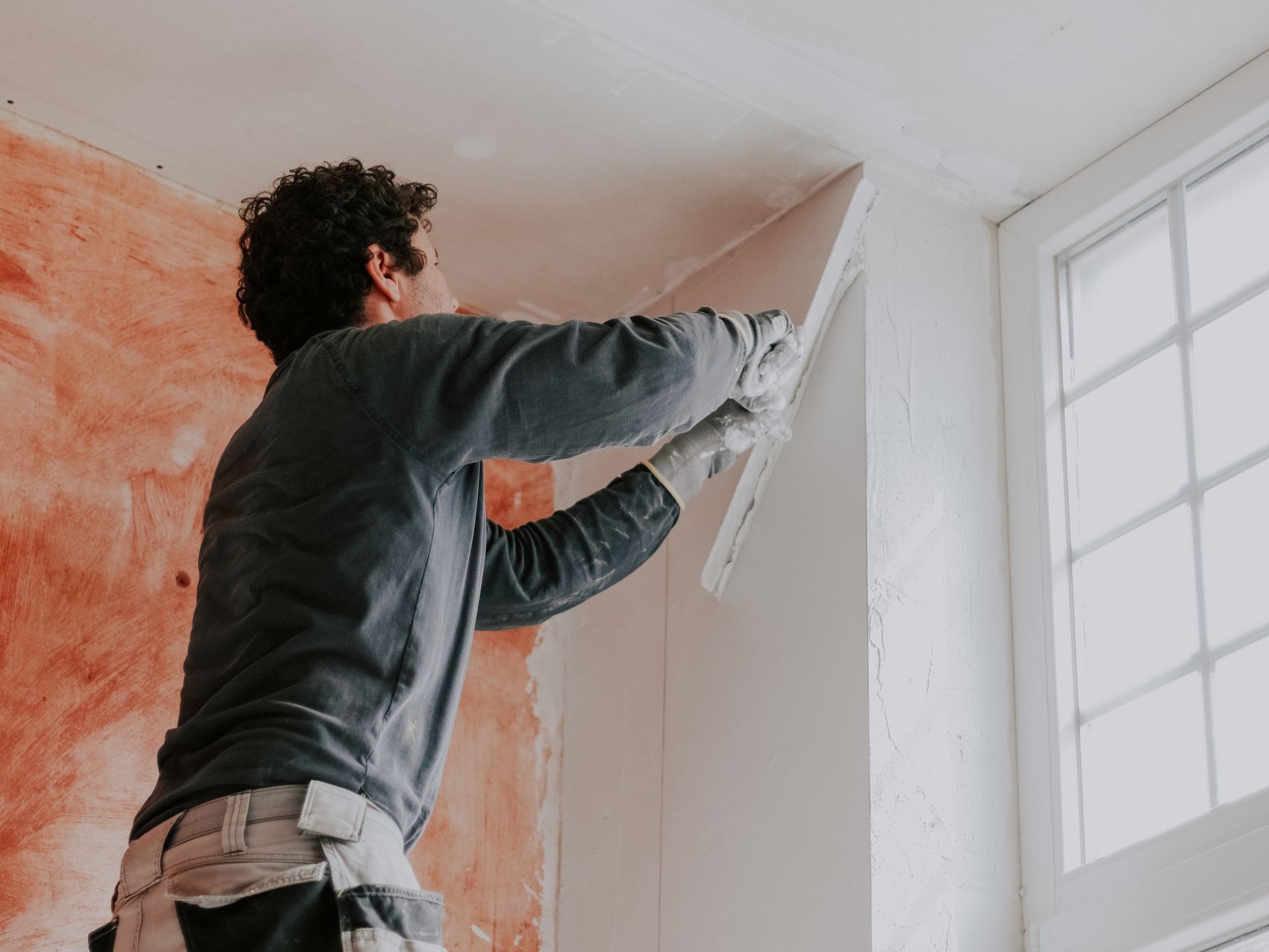 Man smoothing plaster on a wall with a trowel, near a window.