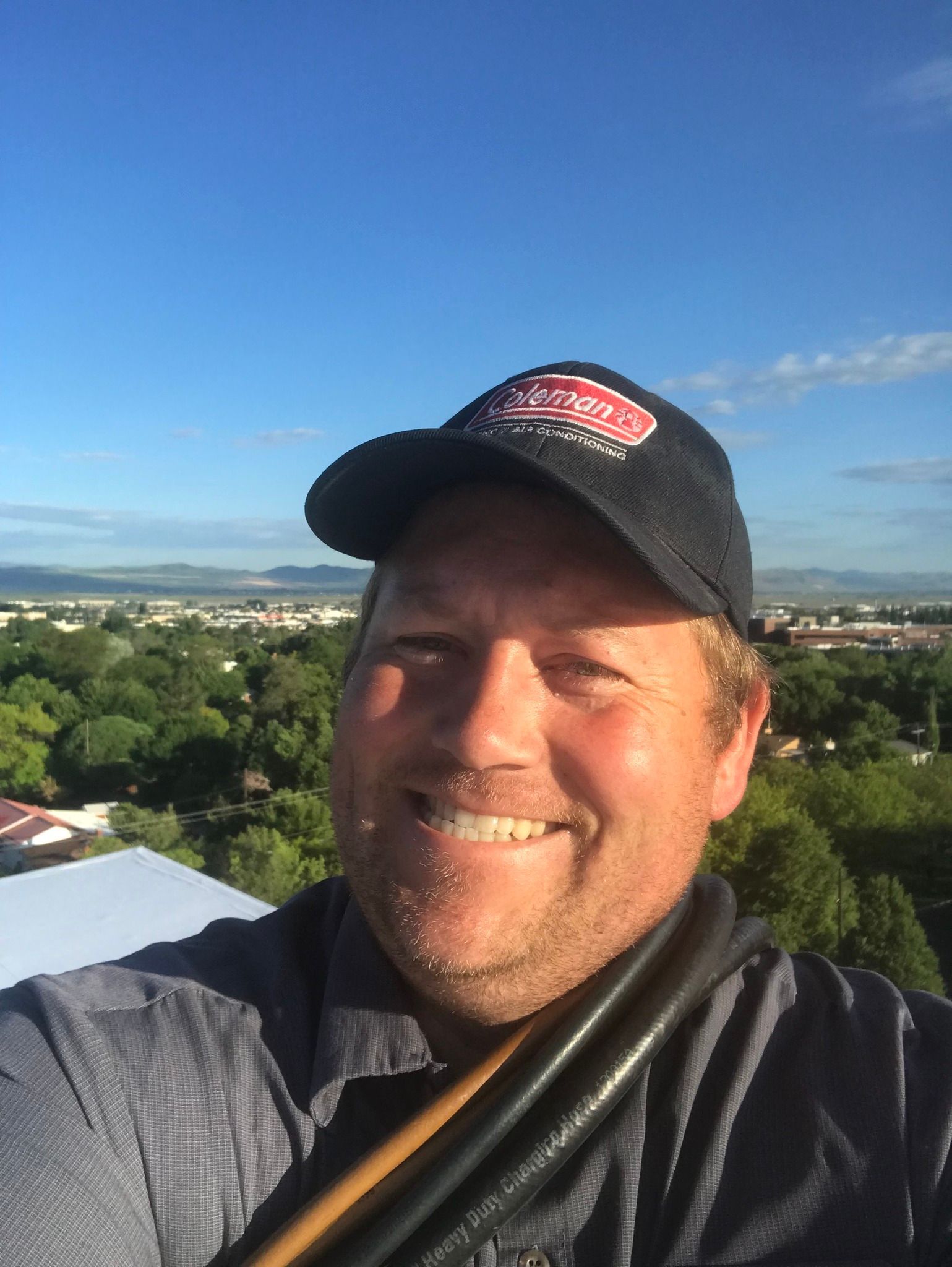 Man in cap smiling, outdoors with city view. Blue sky, green trees.