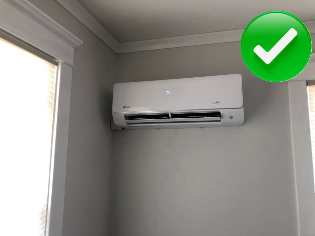 White mini-split AC unit mounted on a gray wall, next to a window. A green checkmark appears in the corner.