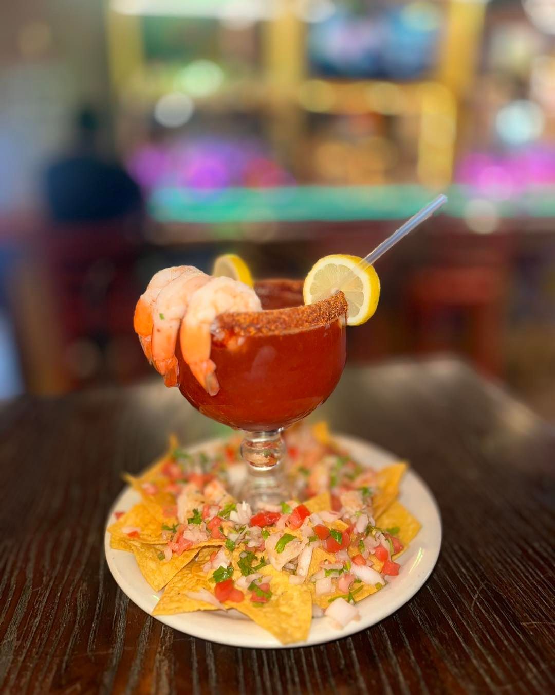Shrimp cocktail in a large glass with nachos on a plate, lemon slice, and a straw.