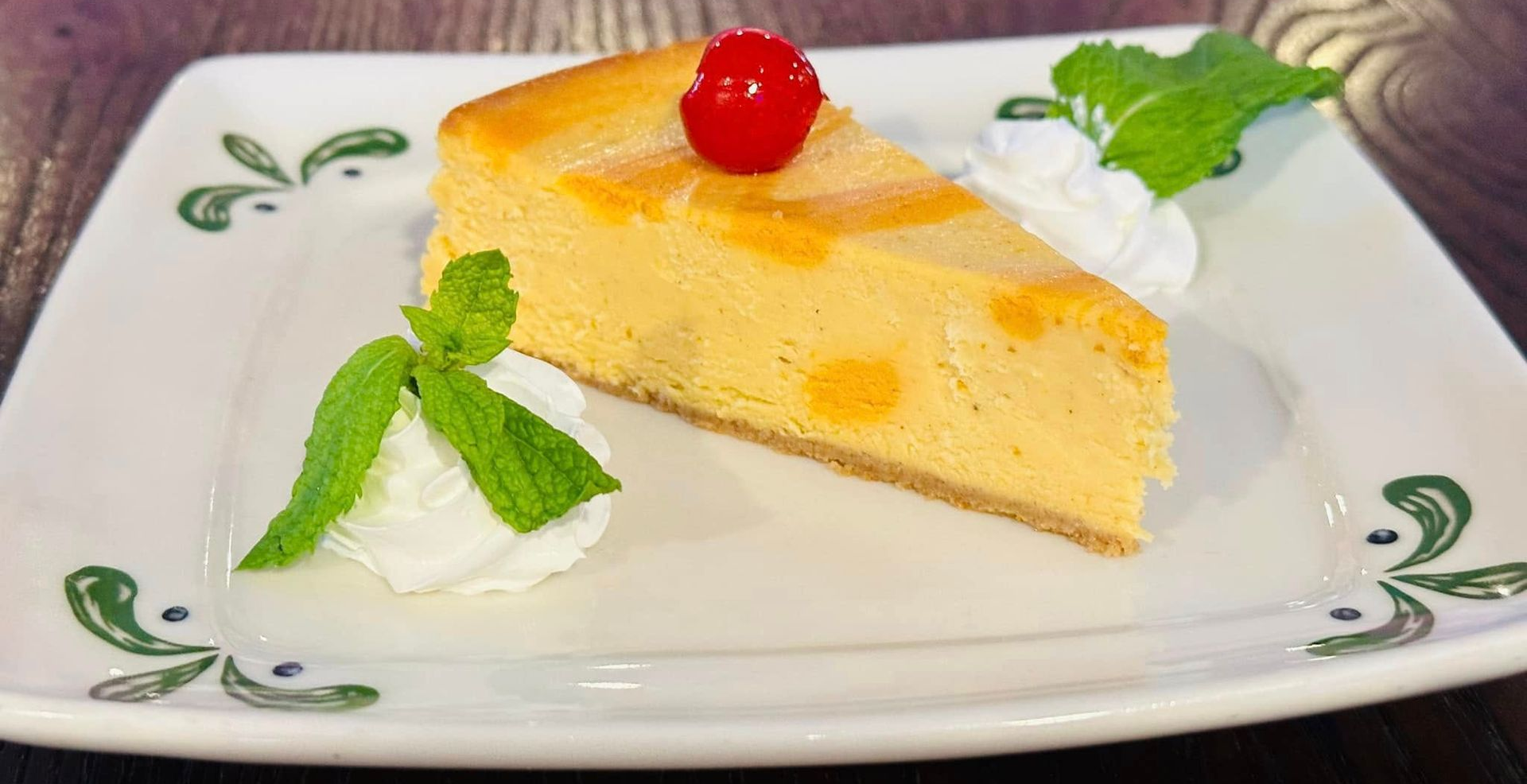 Slice of cheesecake topped with a cherry and mint on a white plate, restaurant setting.