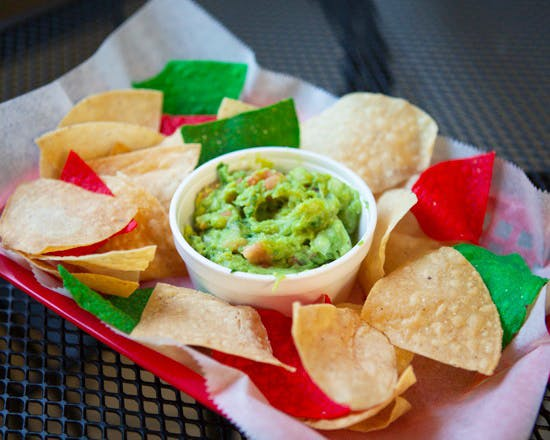 Guacamole dip in a white container surrounded by colorful tortilla chips in a red-lined basket.