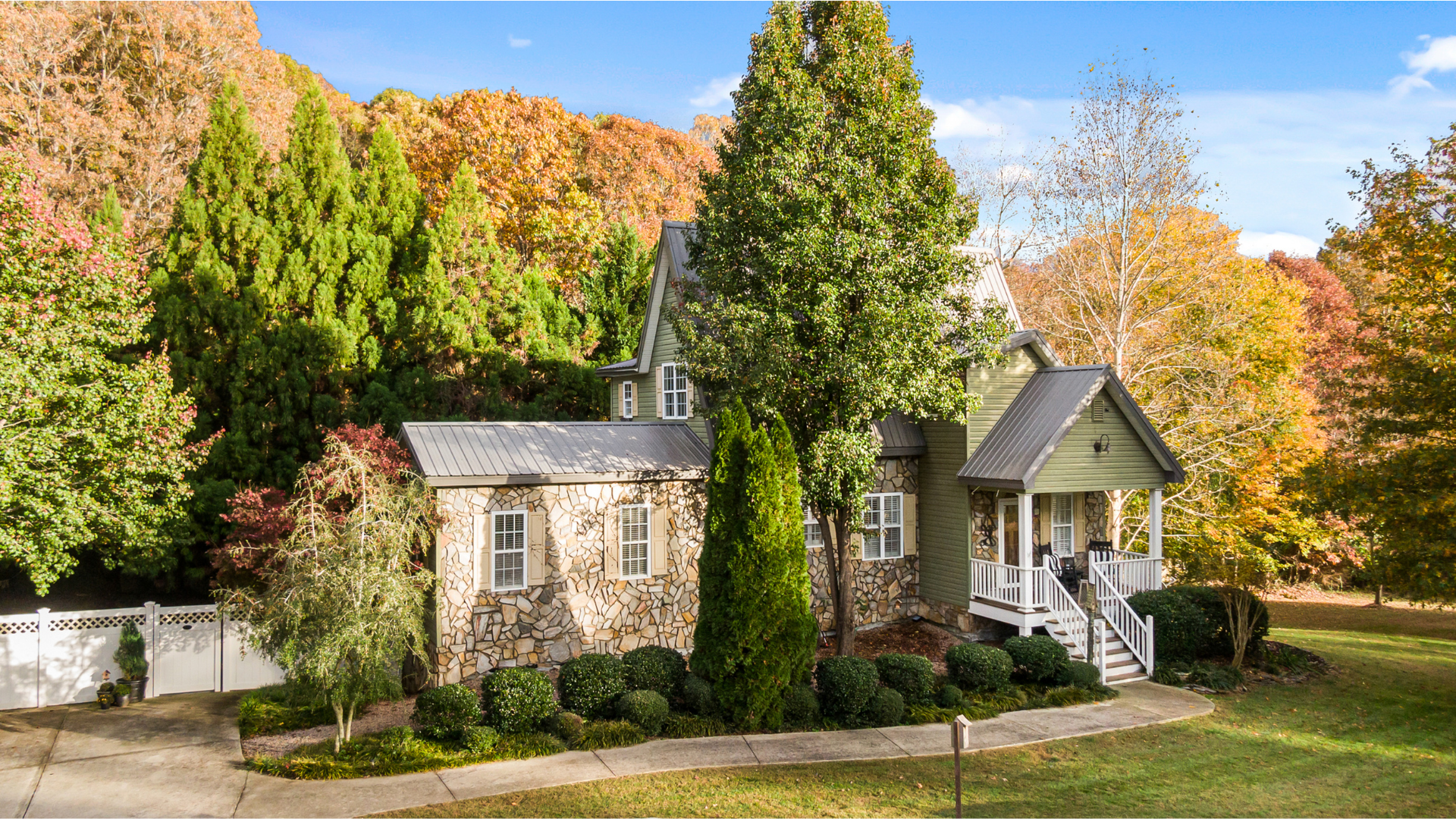 A stone-and-green-sided house with a porch, surrounded by trees with fall foliage.