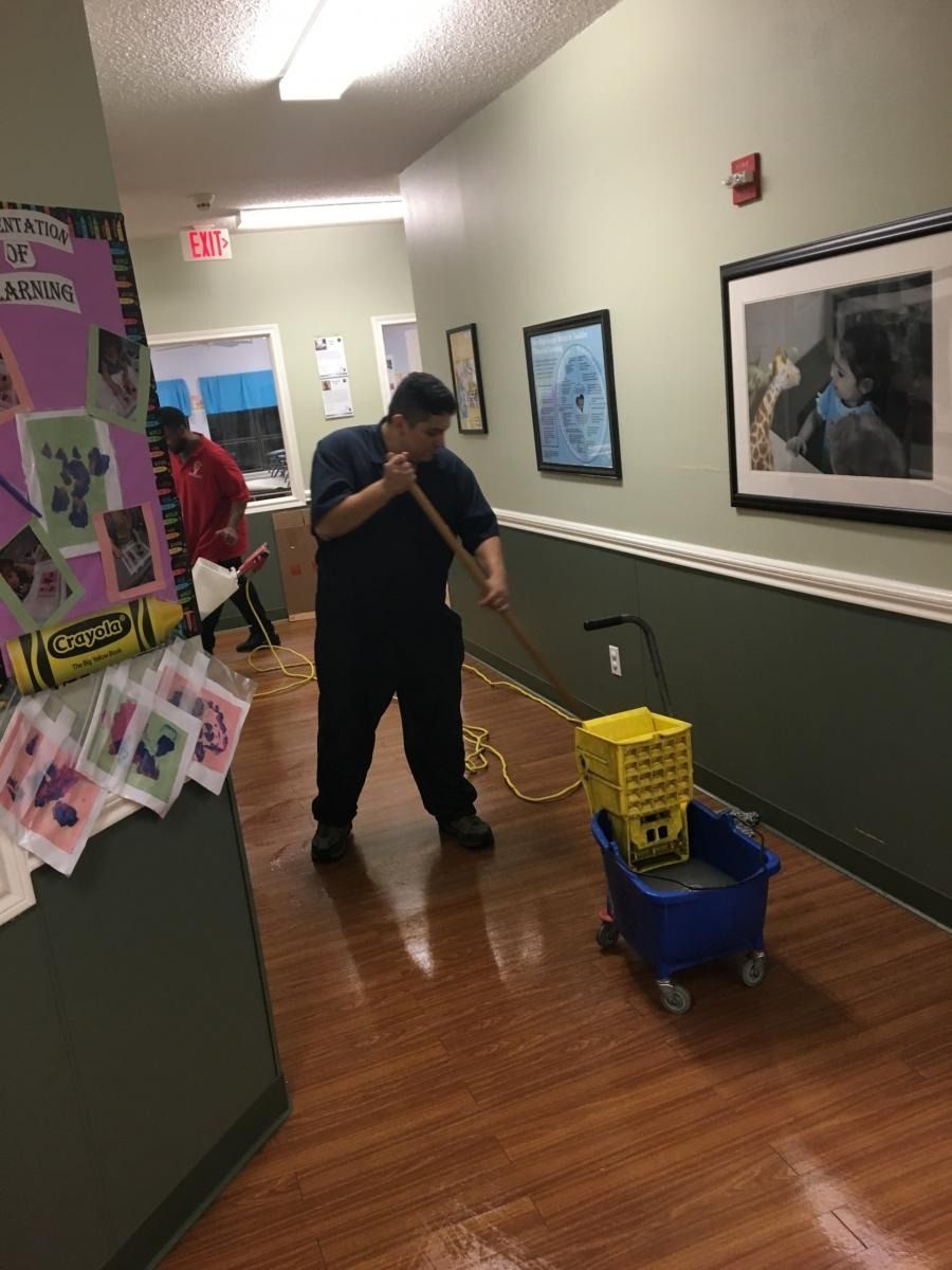 Man mopping a wet hallway floor with a mop and bucket.  Hallway has artwork and a bulletin board.