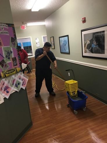 Man mopping a wet hallway floor with a mop and bucket. Hallway has artwork and a bulletin board.