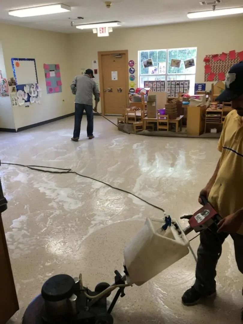 Two people cleaning a classroom floor with a floor cleaner. The room has kid's furniture and decorations.