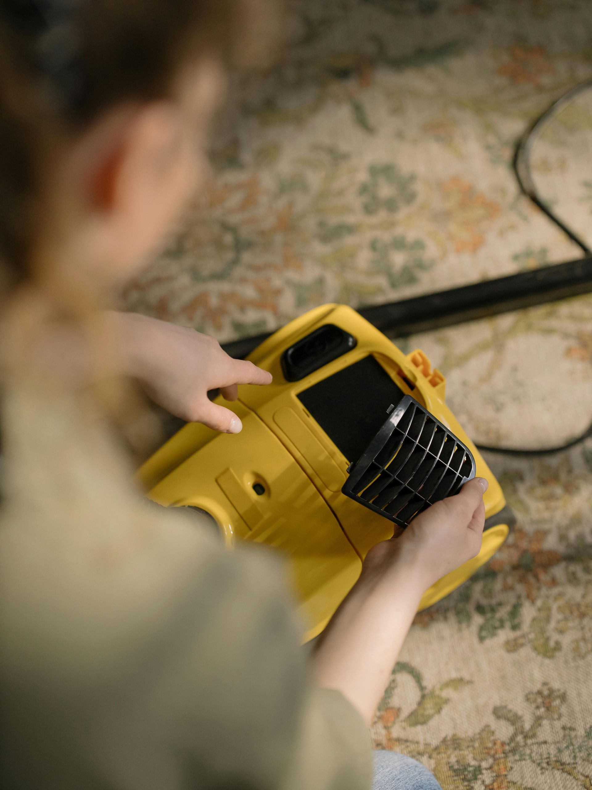 Person removing a filter from a yellow vacuum cleaner on a patterned carpet.