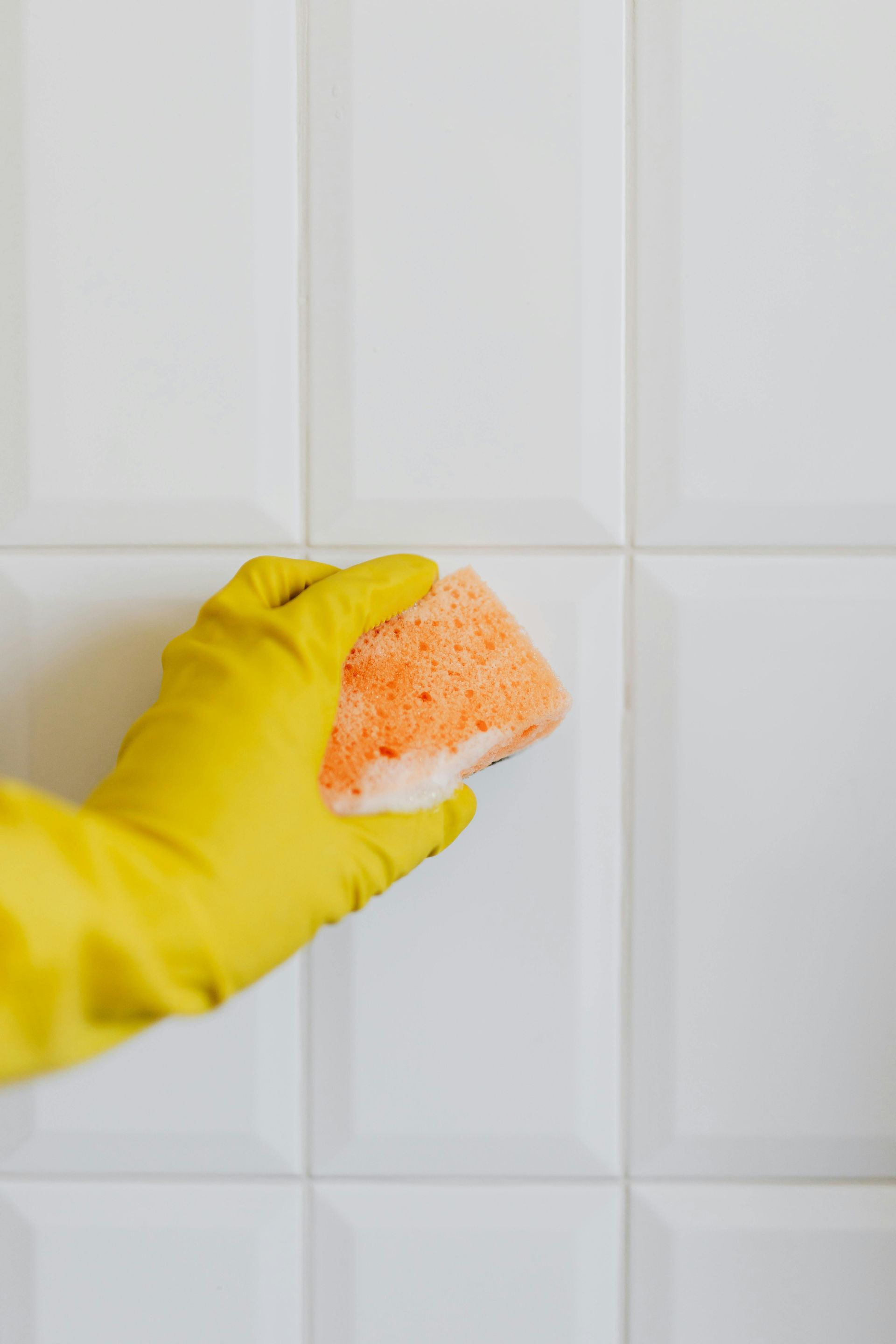Yellow-gloved hand cleaning white tiled wall with an orange sponge.