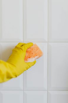 Yellow-gloved hand cleaning white tiled wall with an orange sponge.