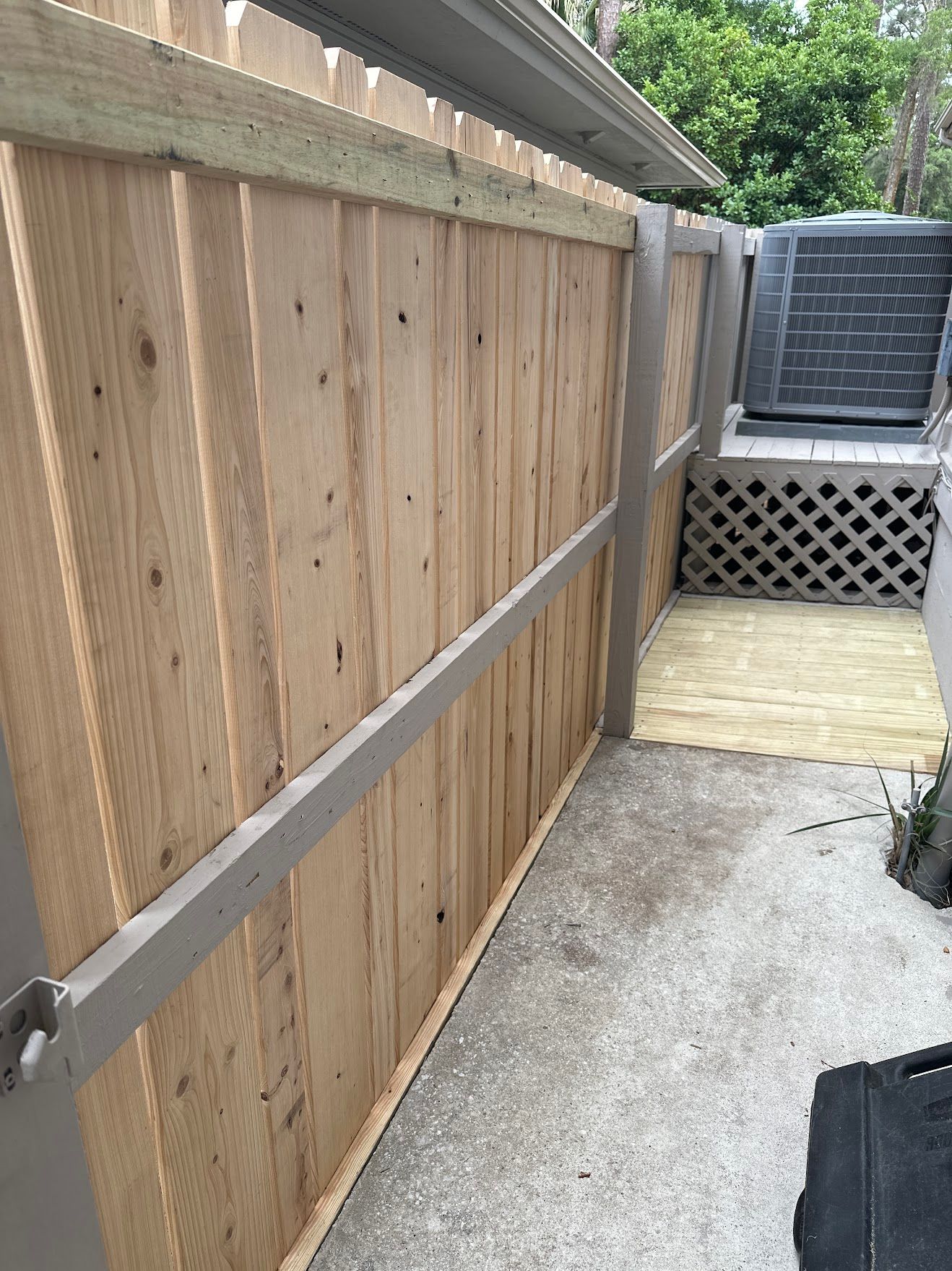 Wooden fence next to a concrete walkway, leading to a small lattice structure and air conditioner.