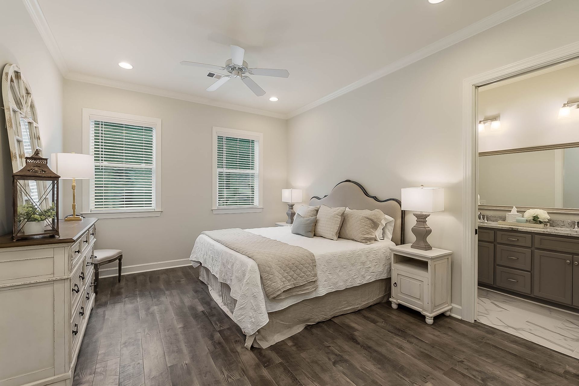 Bedroom with bed, dresser, windows, and en suite bathroom. Light-colored walls and wood-look flooring.